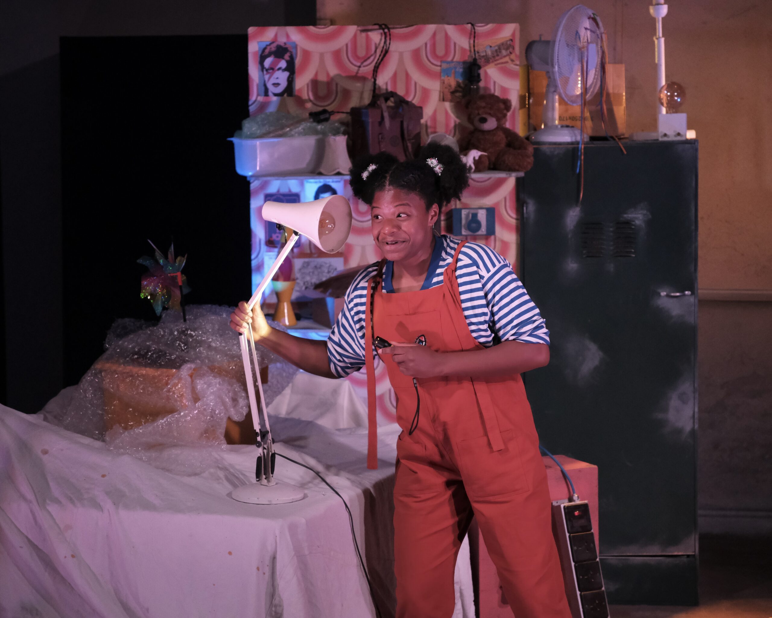 Adrien Spencer, black actor, in orange dungarees, plays with angle poise lamp on set, in character as "Kai"